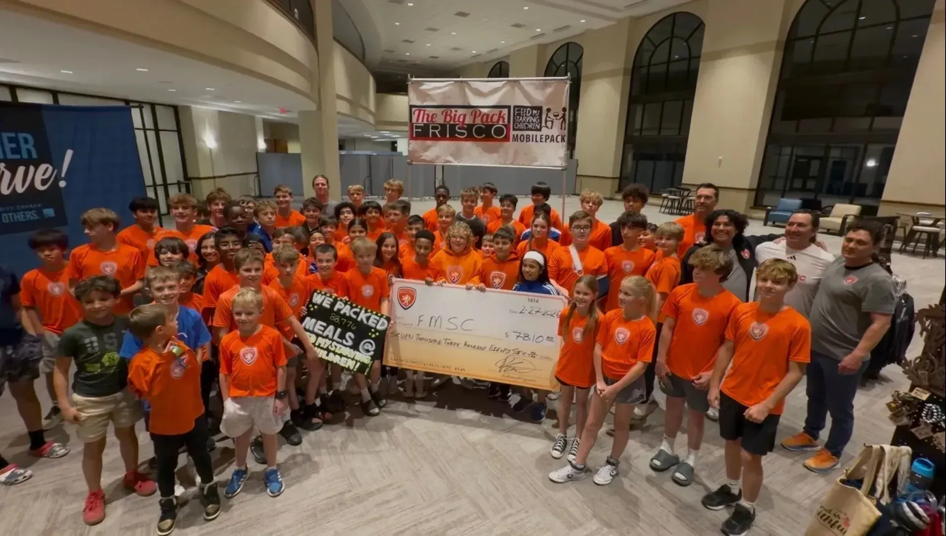 Large group in orange shirts posing indoors with banners at a sports event.