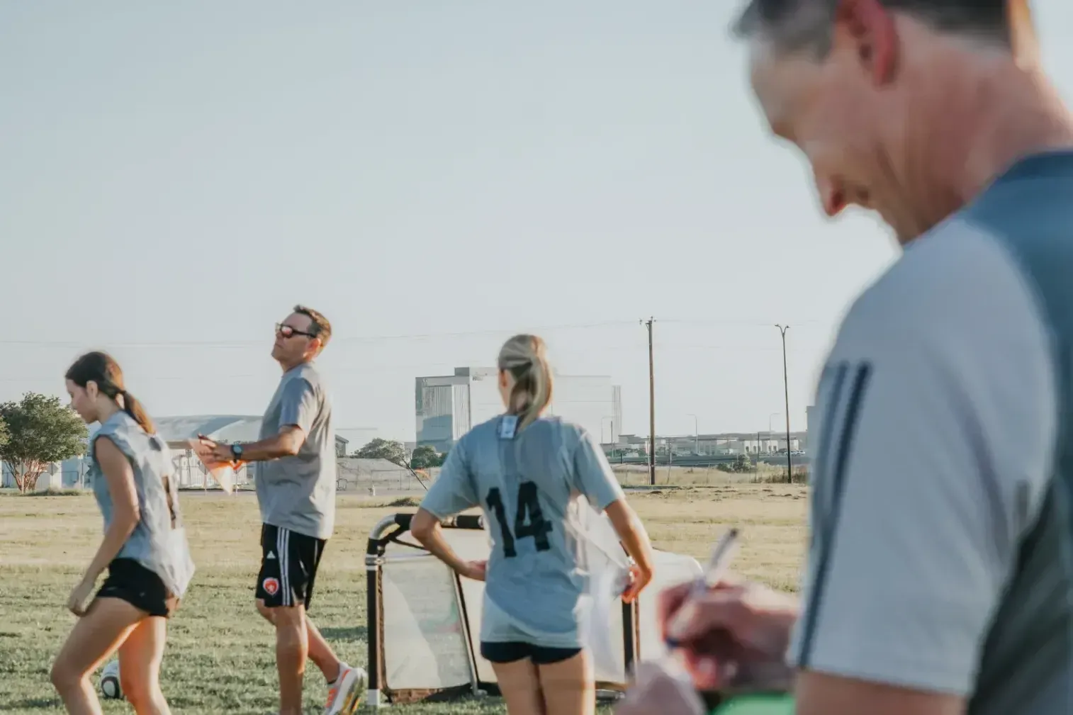 Coach briefing a soccer team on a field, with players holding clipboards around him.