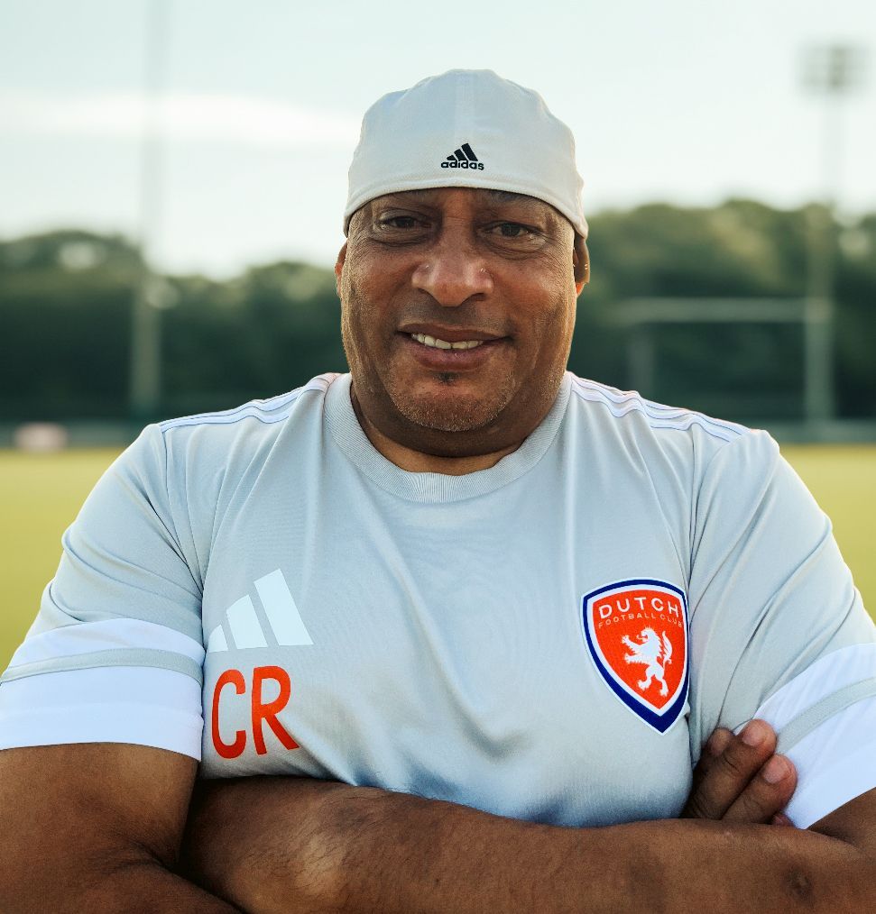 Soccer coach in white shirt speaking on a grassy field with players in orange jerseys behind him