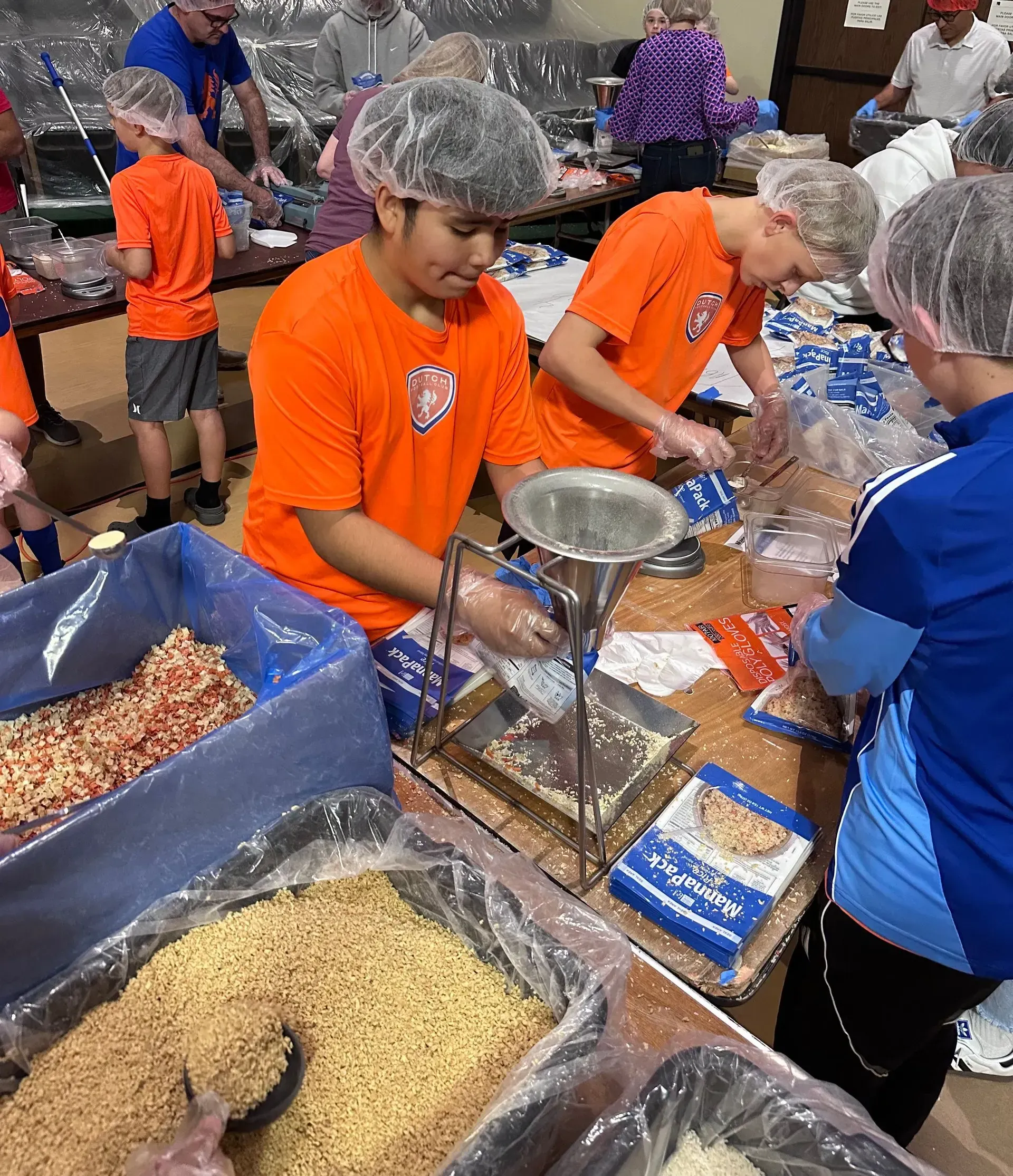Volunteers in orange shirts scooping food into trays at a crowded packing station