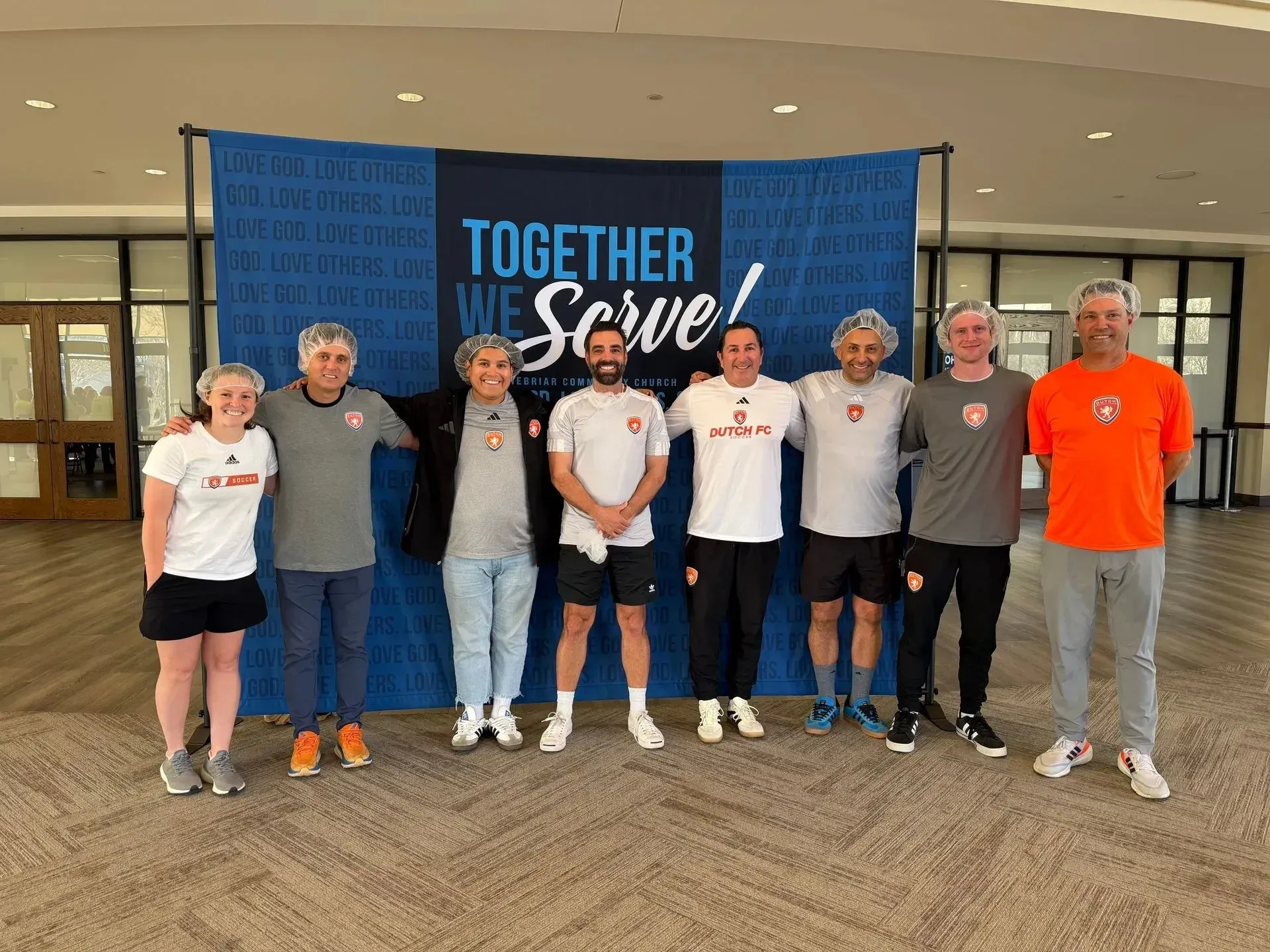 Seven people standing in front of a blue “Together We Serve” banner inside a lobby