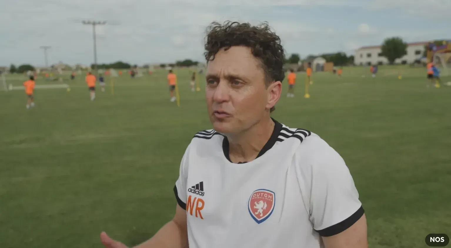 Soccer coach in white shirt speaking on a grassy field with players in orange jerseys behind him