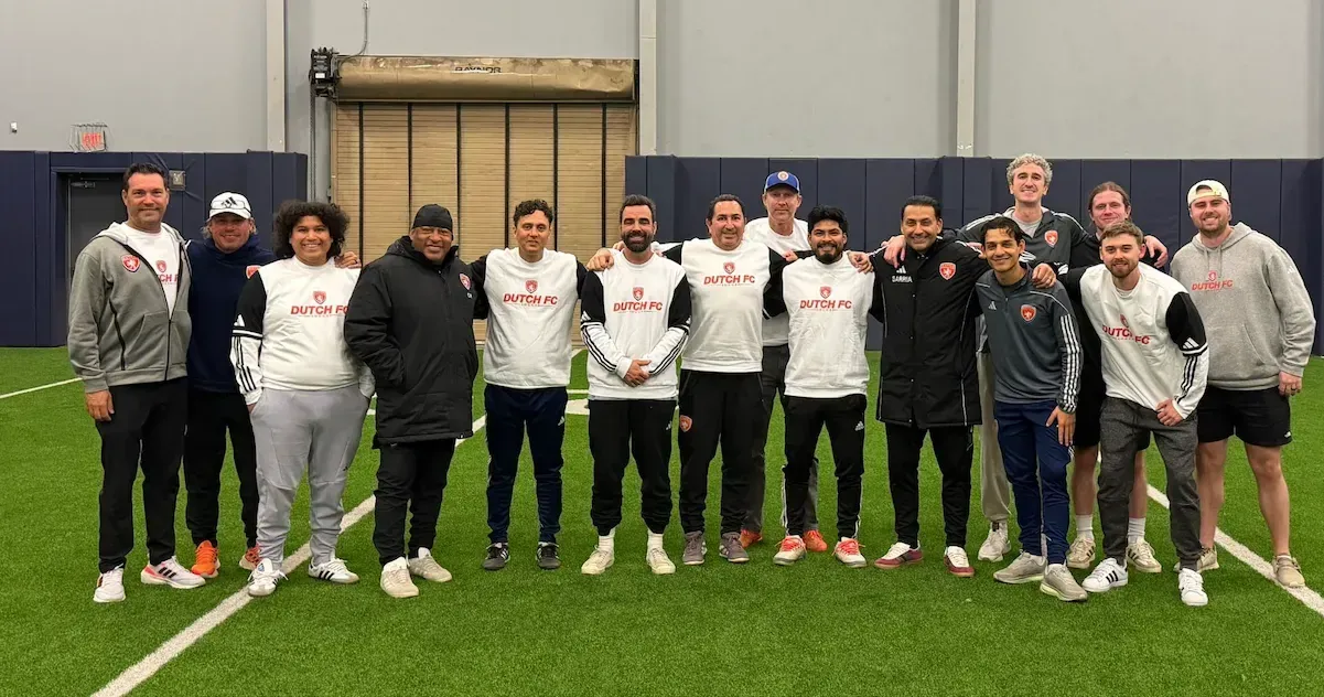 Soccer team posing on an indoor turf field, wearing white and black uniforms, with a goal behind them