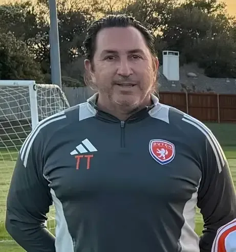 Soccer coach in white shirt speaking on a grassy field with players in orange jerseys behind him