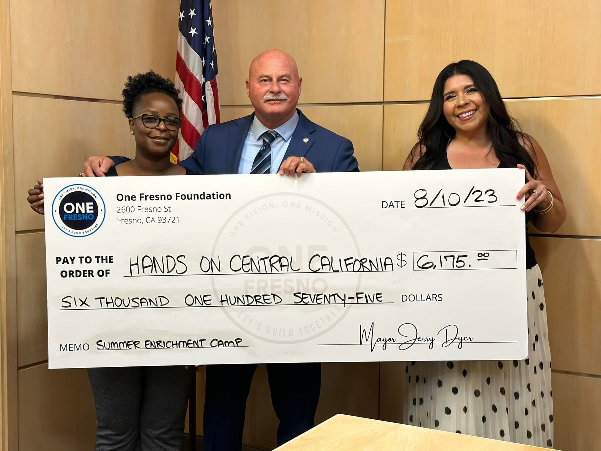A man and two women are holding a large check in front of an american flag.