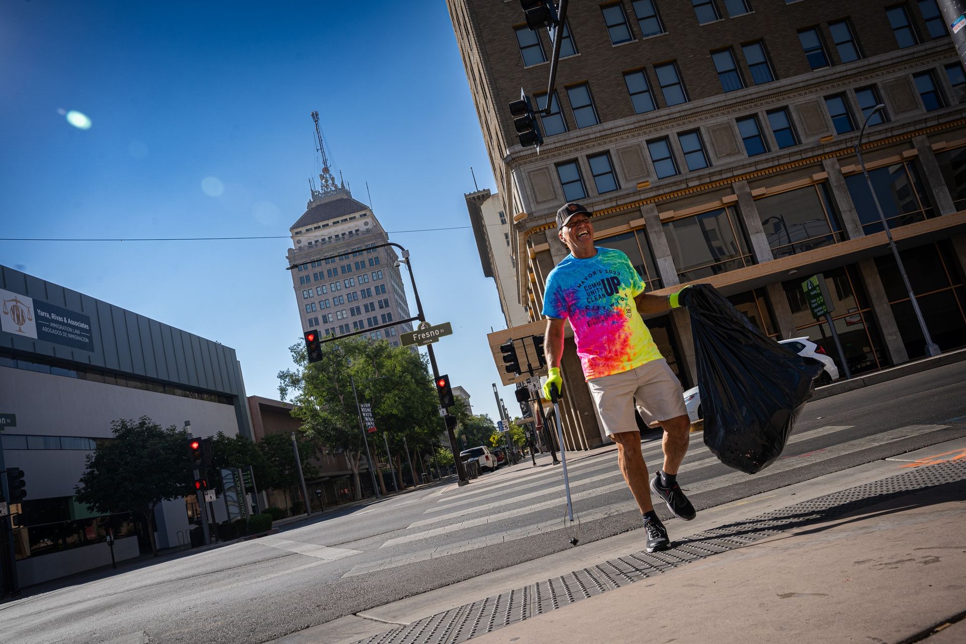 A man in a tie dye shirt is walking down the street with a trash bag.
