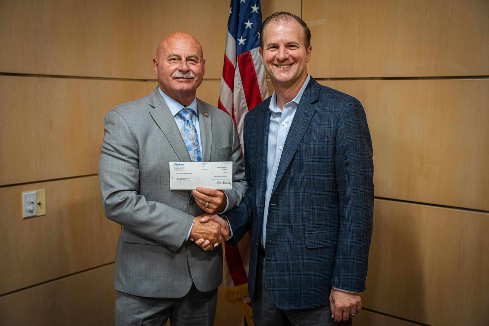 Two men are shaking hands while holding a check in front of an american flag.