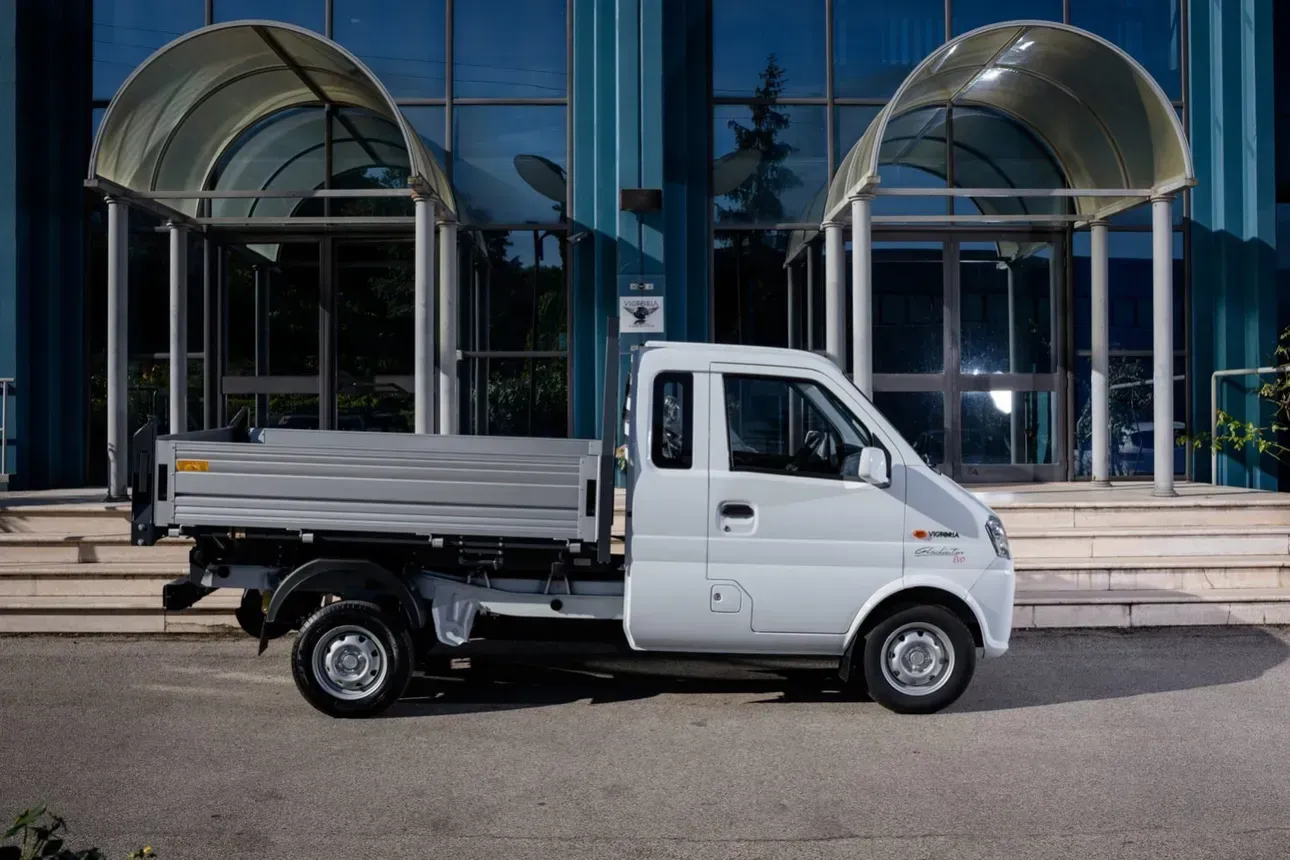 Mini camioncino bianco con pianale parcheggiato davanti a un edificio con porte a vetri e tende da sole ad arco