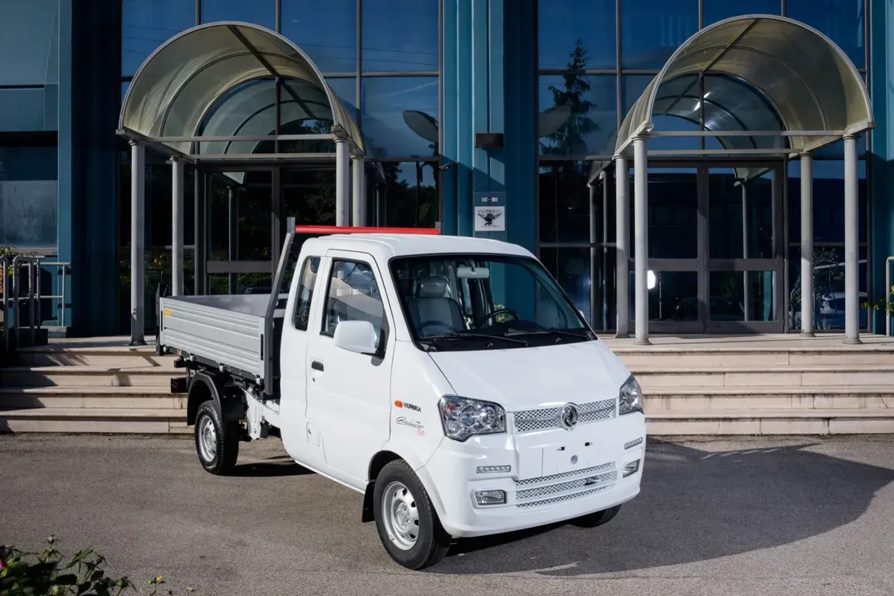 Mini dumper bianco parcheggiato davanti a un edificio in vetro con ingressi ad arco.
