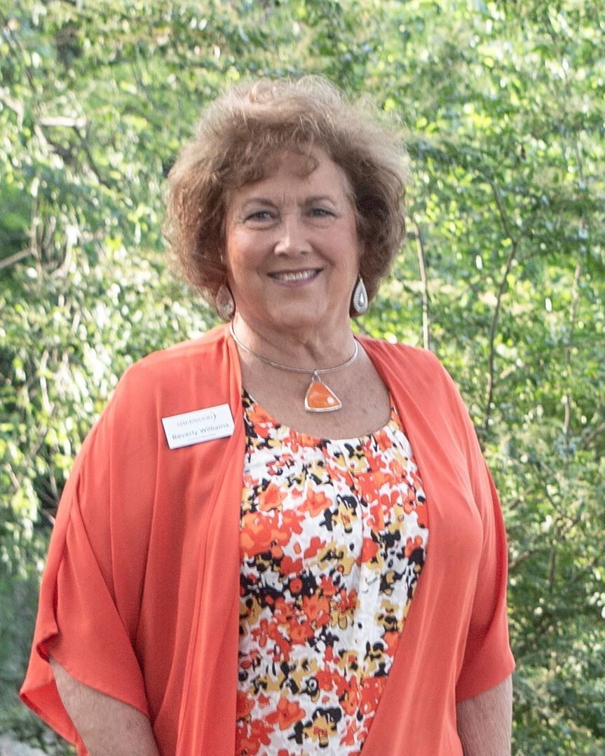 Woman with curly brown hair, smiling, wearing orange cardigan over floral top, standing outdoors.