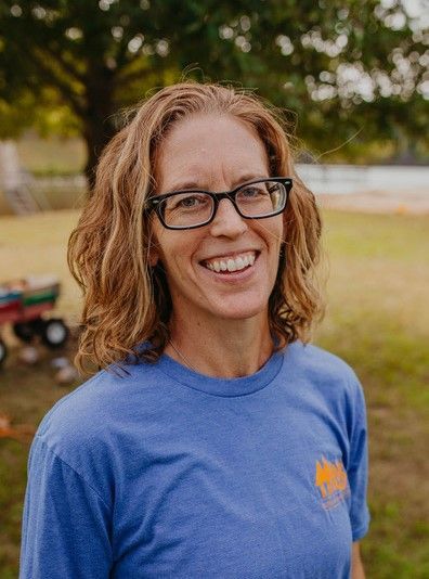 Woman with glasses smiling, blue shirt, outdoors in front of a tree and red wagon.