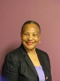 Smiling Black woman in a black blazer, purple top, and gold jewelry against a pink background.