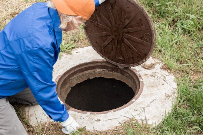 Worker in blue jacket opening a round manhole cover over a dark sewer opening.
