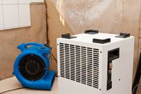 A blue industrial floor dryer sits next to a white commercial dehumidifier in a room with water-damaged walls.
