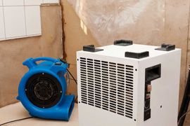 Blue fan and white dehumidifier set up in a room with exposed wall.