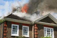 A brick house with flames and thick smoke billowing from its roofline against a cloudy sky.