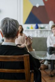 A person with grey hair sits in a wooden chair, holding a pen, facing two other people in a light, modern interior.