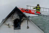 A firefighter in protective gear works on the roof of a house with fire damage near an upper-level window.