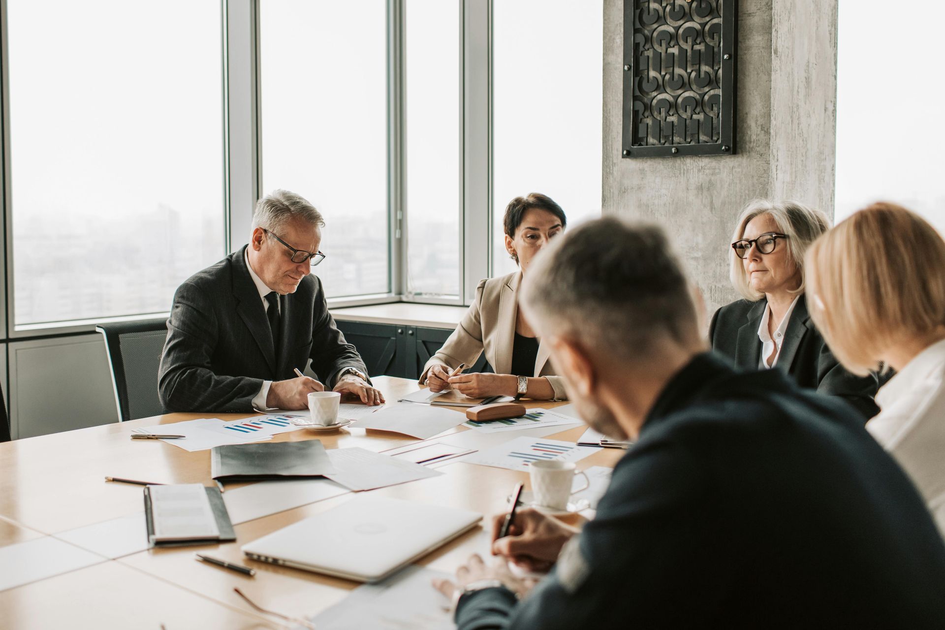 People in business attire at a conference table, reviewing documents, with a city view.