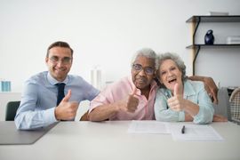 Three people at a table giving thumbs up; one is wearing a suit, two are older adults.