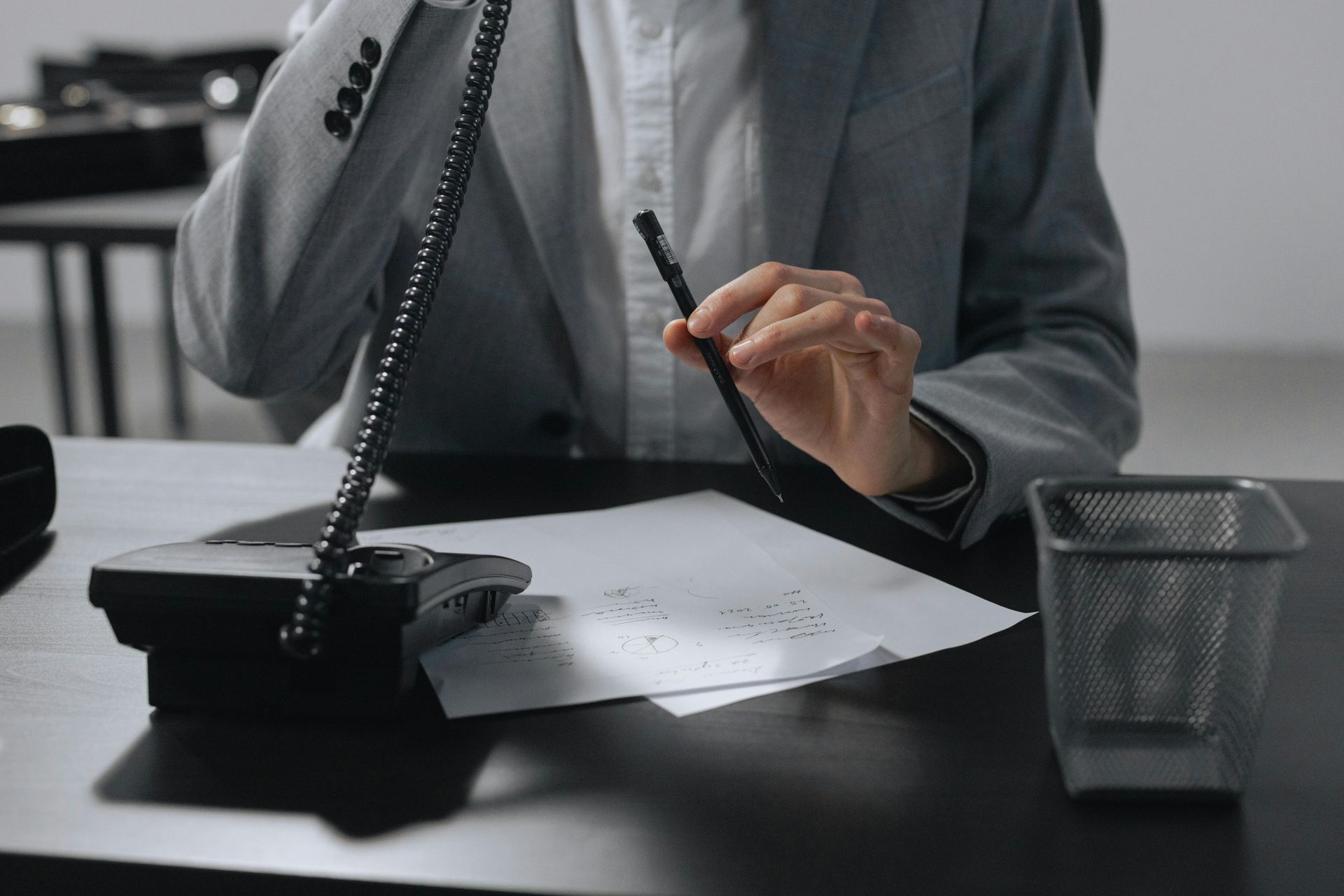A person in a gray suit holding a pen over paper while talking on a telephone at a desk with a wire mesh bin.