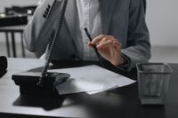 A person in a gray suit holding a pen over paper while talking on a telephone at a desk with a wire mesh bin.