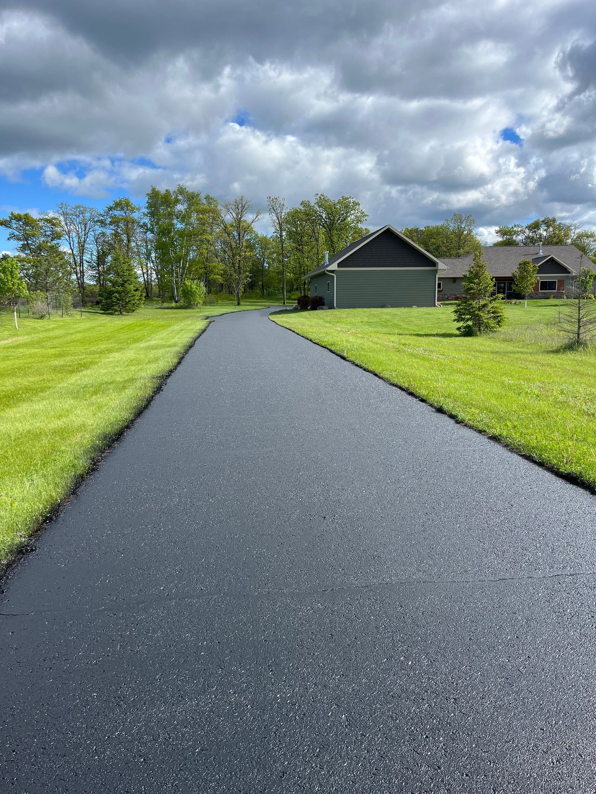 A black asphalt driveway leading to a house surrounded by green grass.