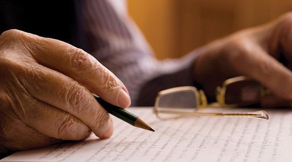 Close-up of elderly person's hand holding pencil, writing on paper; eyeglasses rest nearby.