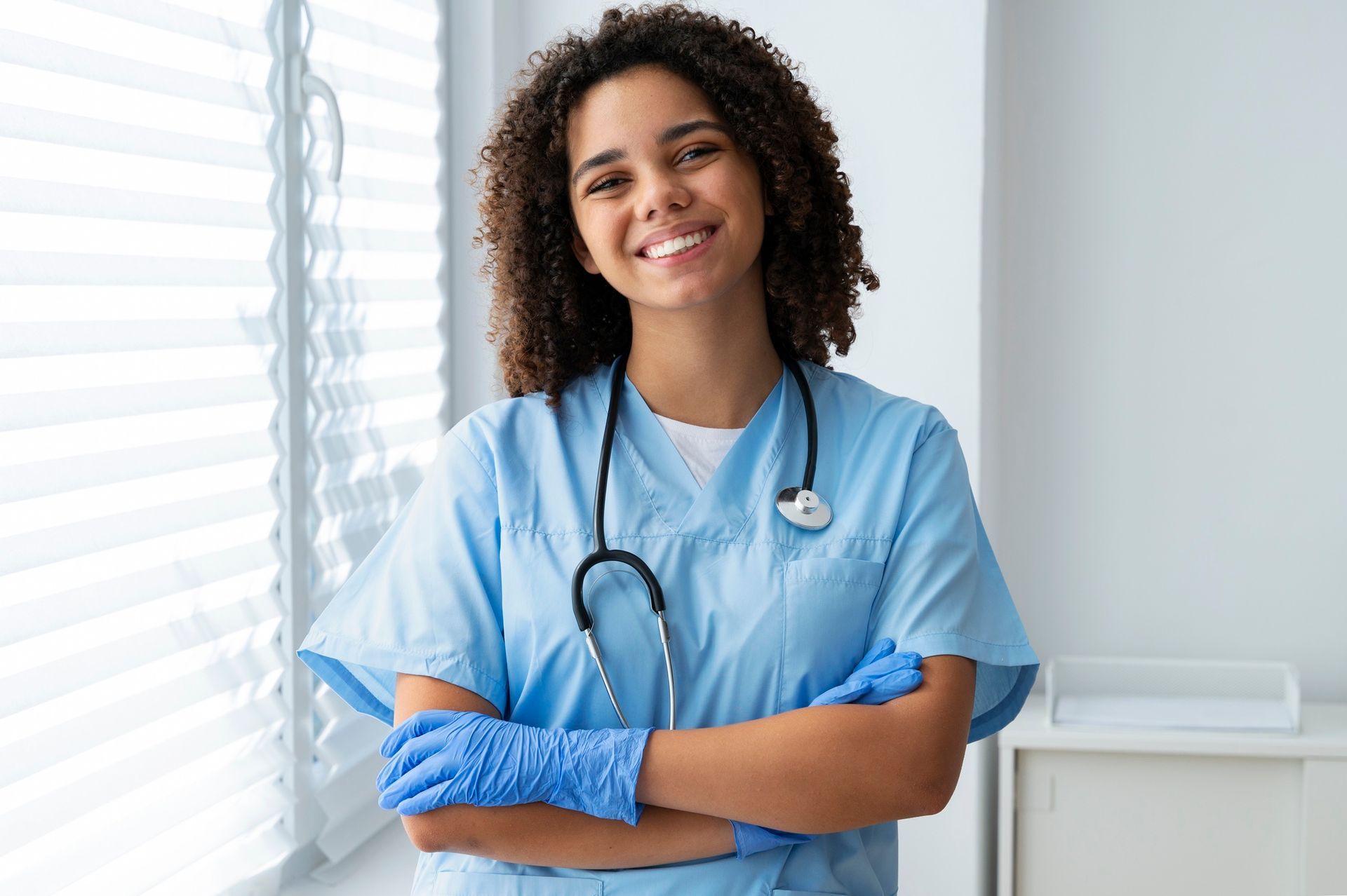 Smiling healthcare worker in blue scrubs and gloves, arms crossed, standing by a bright window