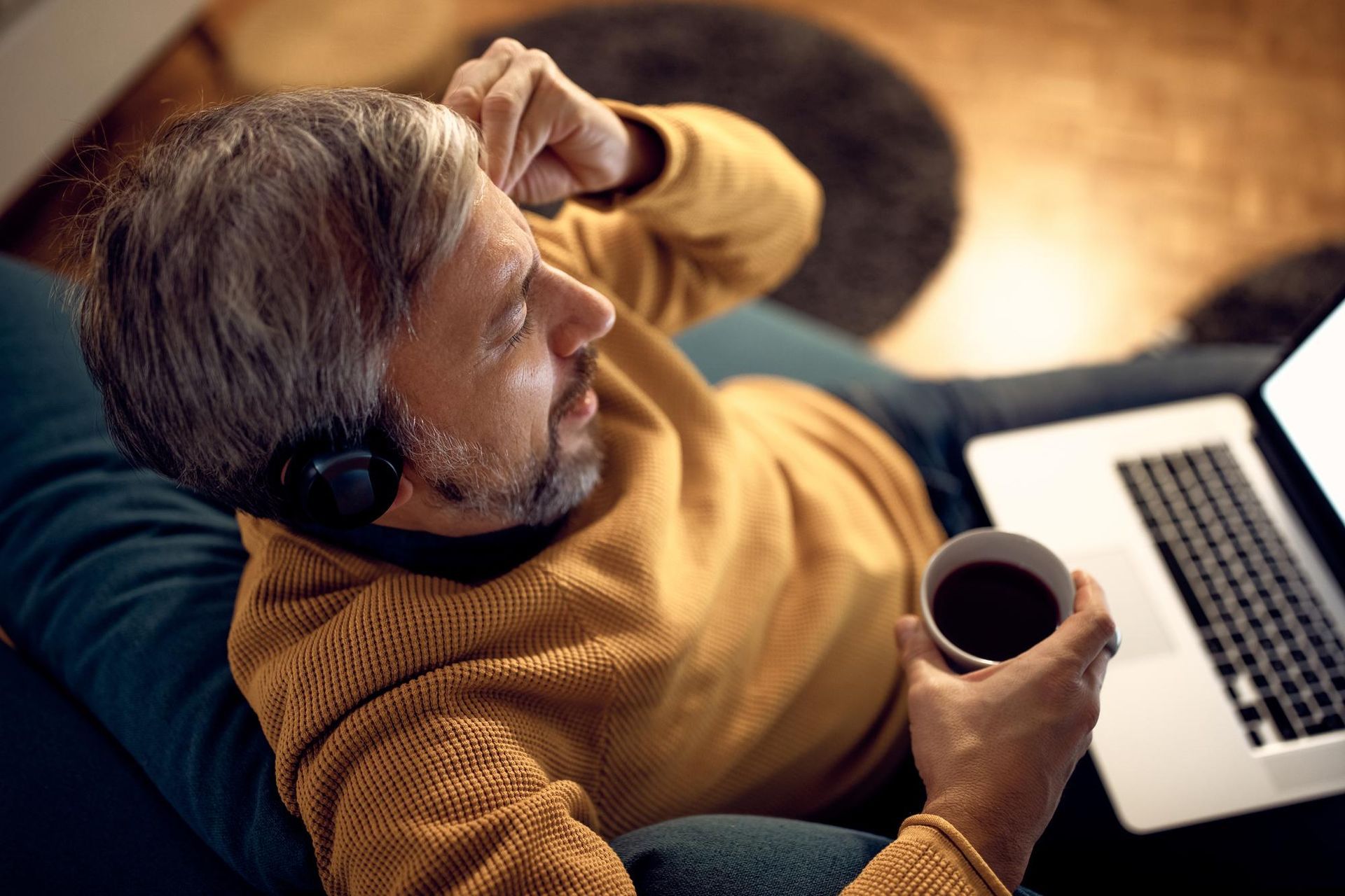 Person reclining on a couch with a laptop and coffee mug, wearing a mustard sweater