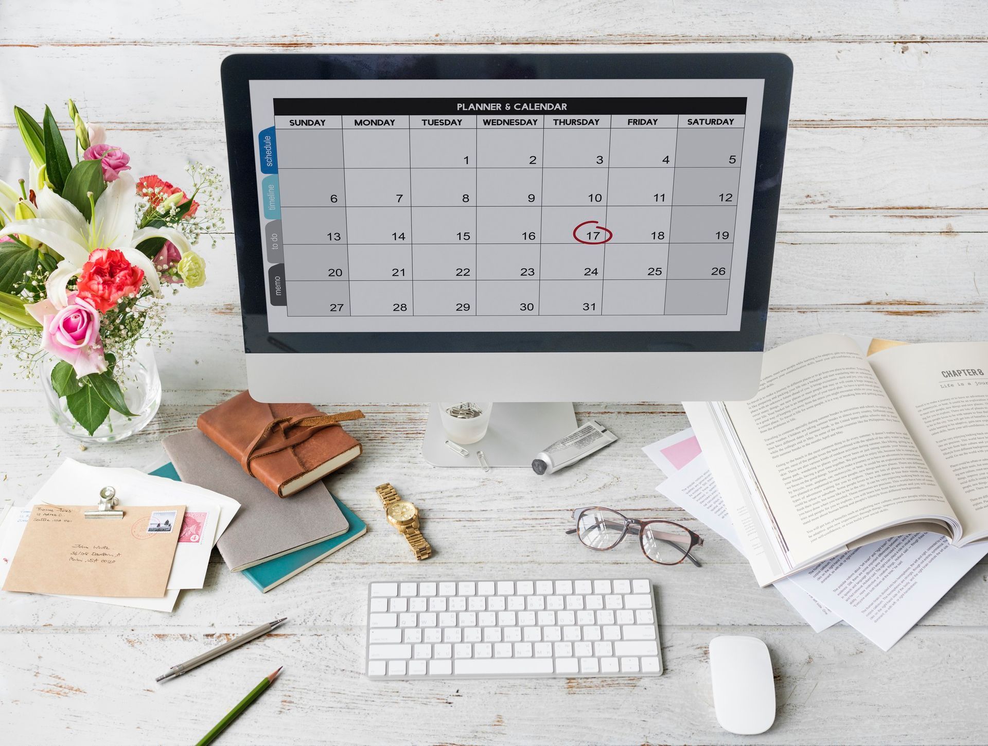 Desktop workspace with calendar on monitor, keyboard, mouse, flowers, notebooks, and papers on a white desk