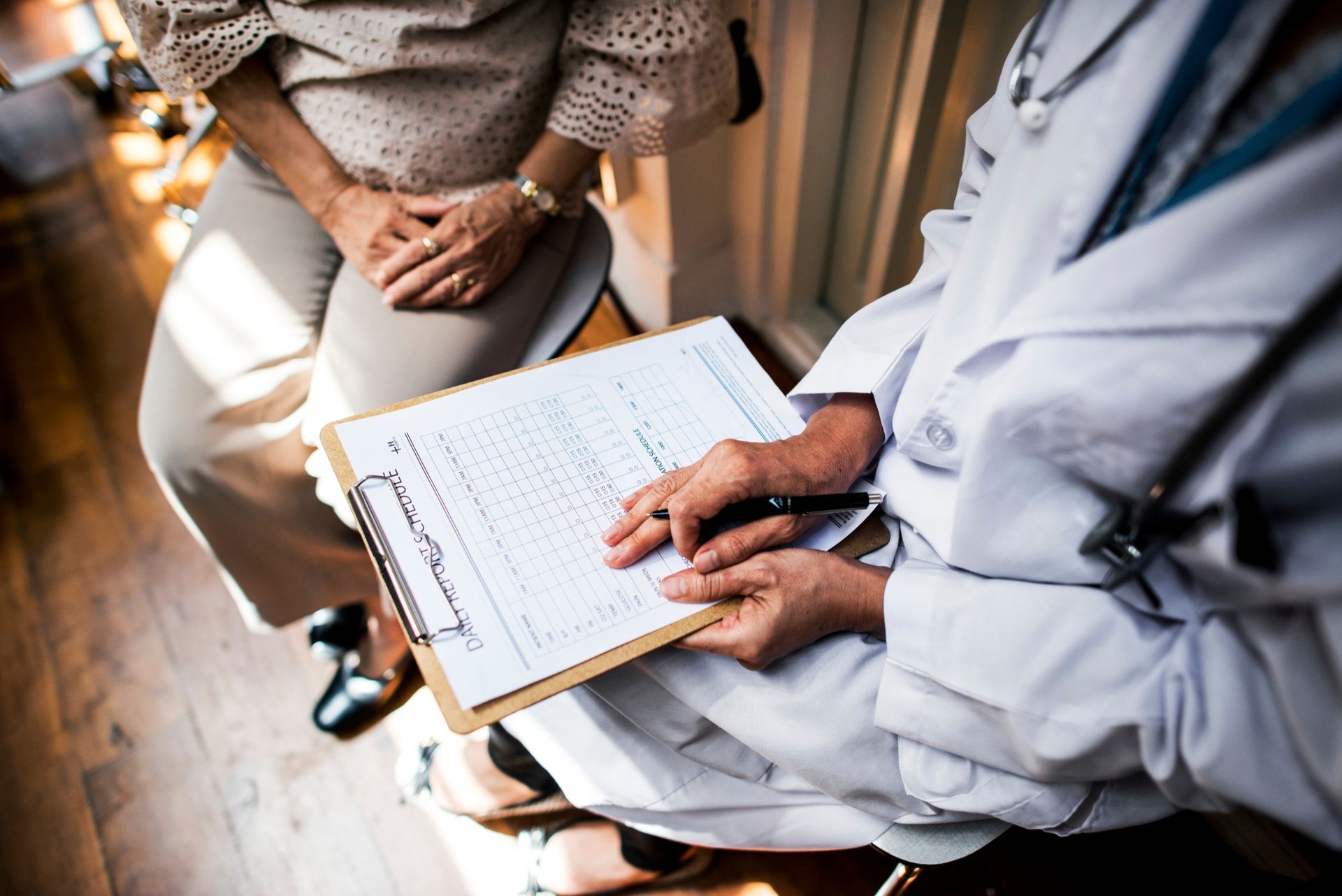Doctor holding a clipboard and writing notes beside a seated patient in a clinic