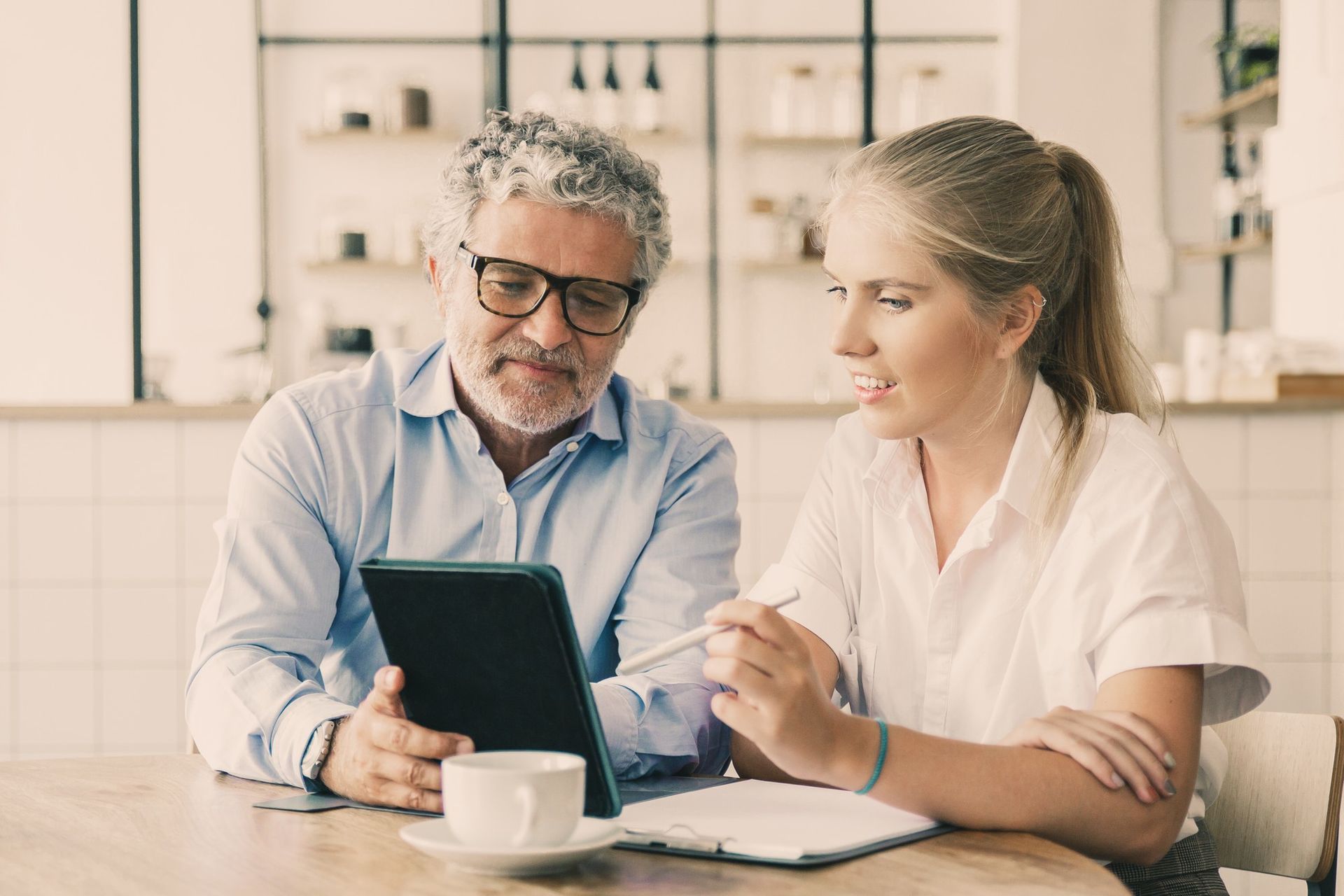 Two people reviewing a tablet at a table with a notebook and coffee in a bright office