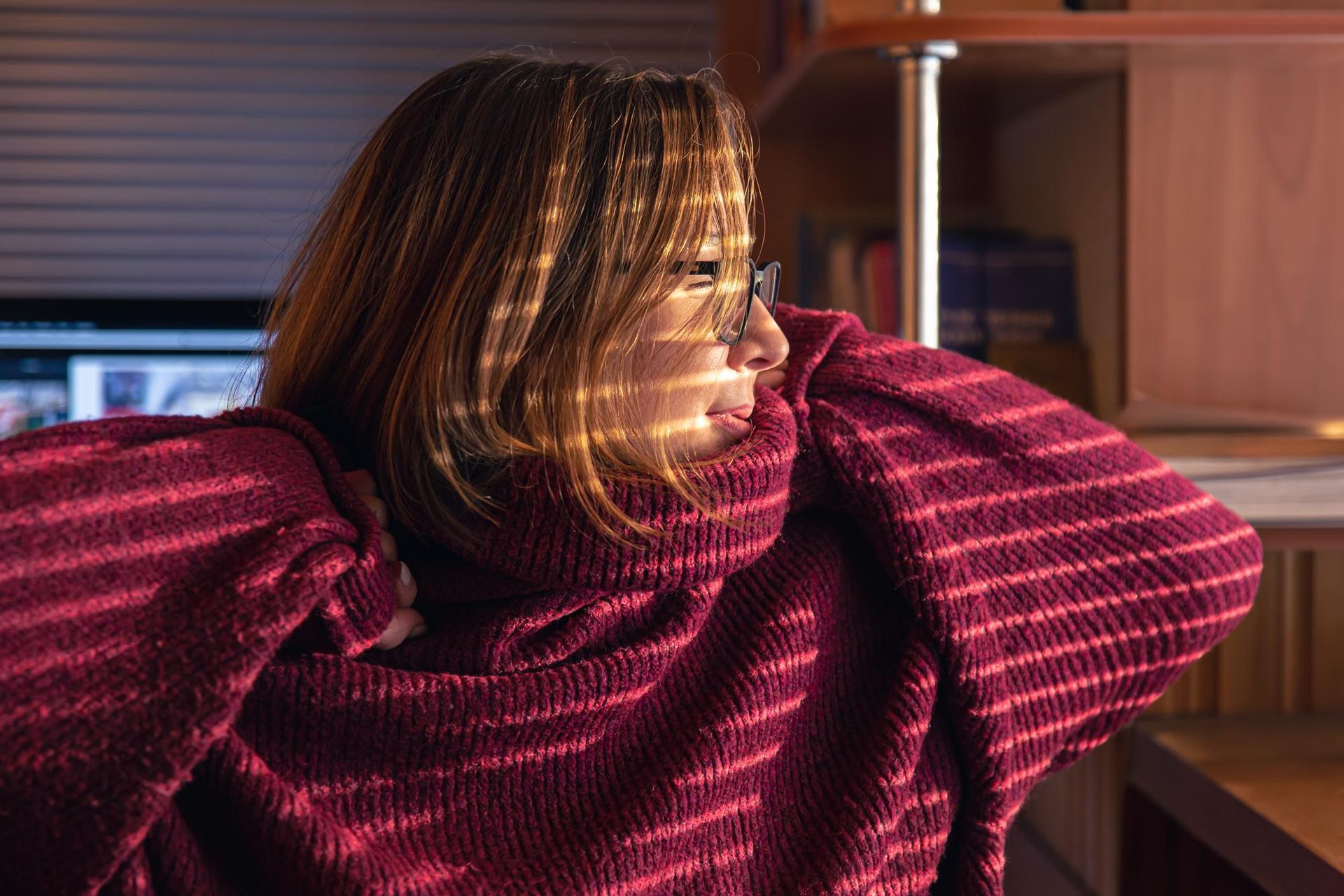 Person in a red sweater leaning by a window, lit by sunlight with striped shadows across their face.