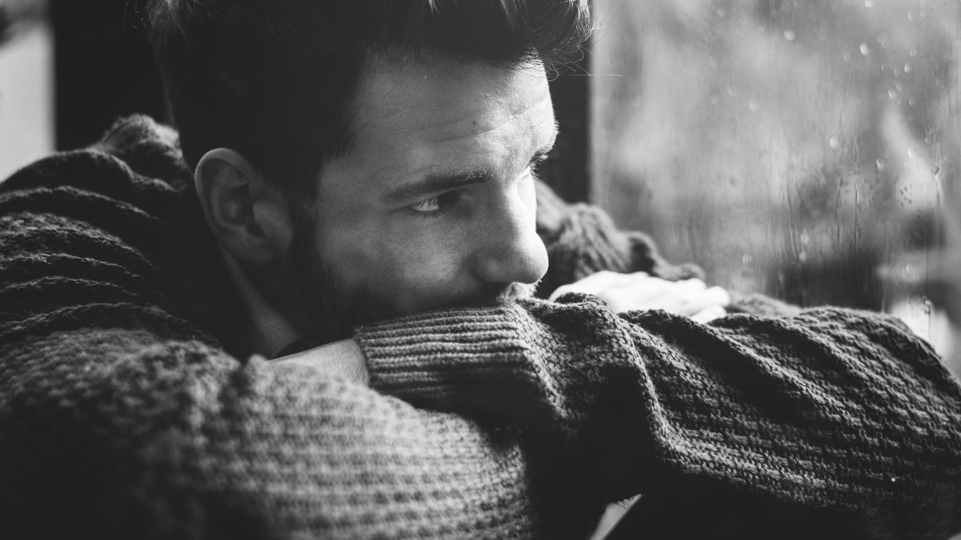 A man in black and white rests his head on folded arms, looking solemn outdoors.