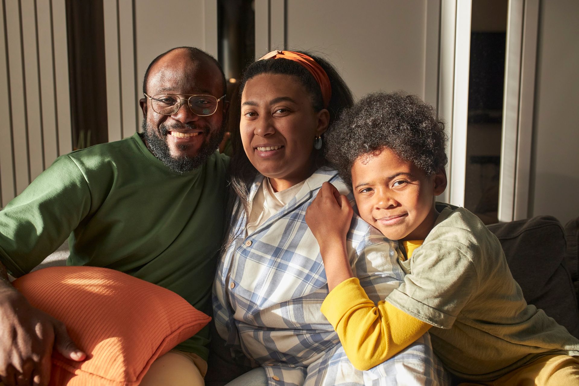 Three people sitting closely on a couch, smiling in warm sunlight.