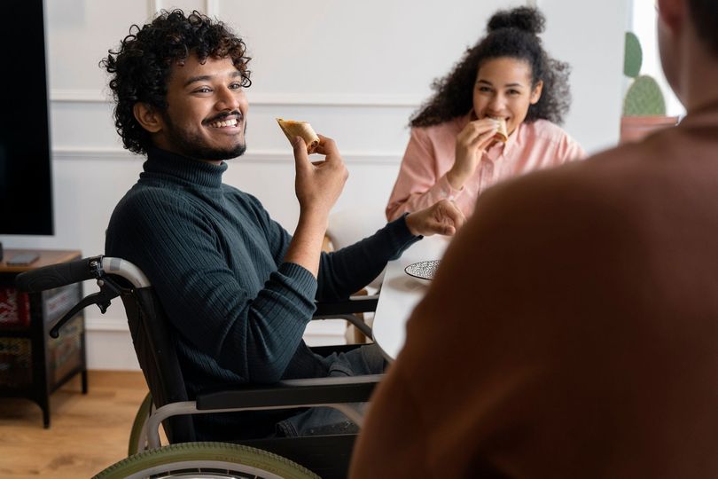Smiling person in a wheelchair holding a snack during a casual meeting with others indoors
