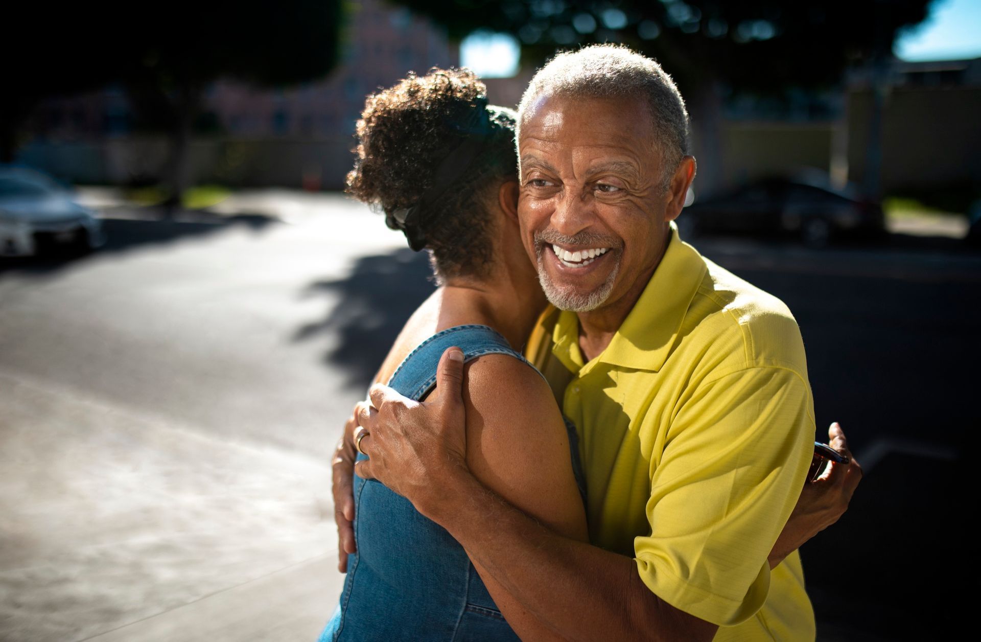 Two people hugging outdoors, one smiling in a yellow shirt on a sunlit street.