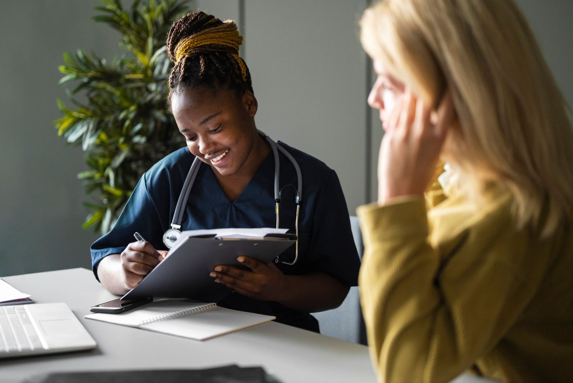 Doctor writing on a clipboard while speaking with a seated person at a desk in a bright office