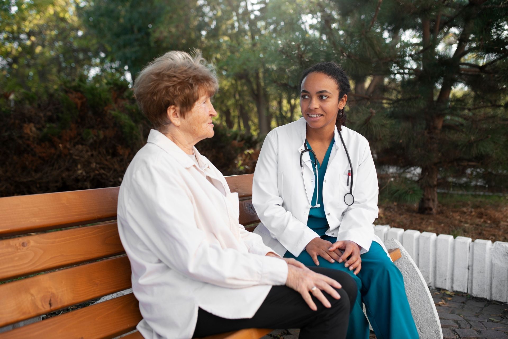 Two people in white coats talk on a park bench, one holding a stethoscope.
