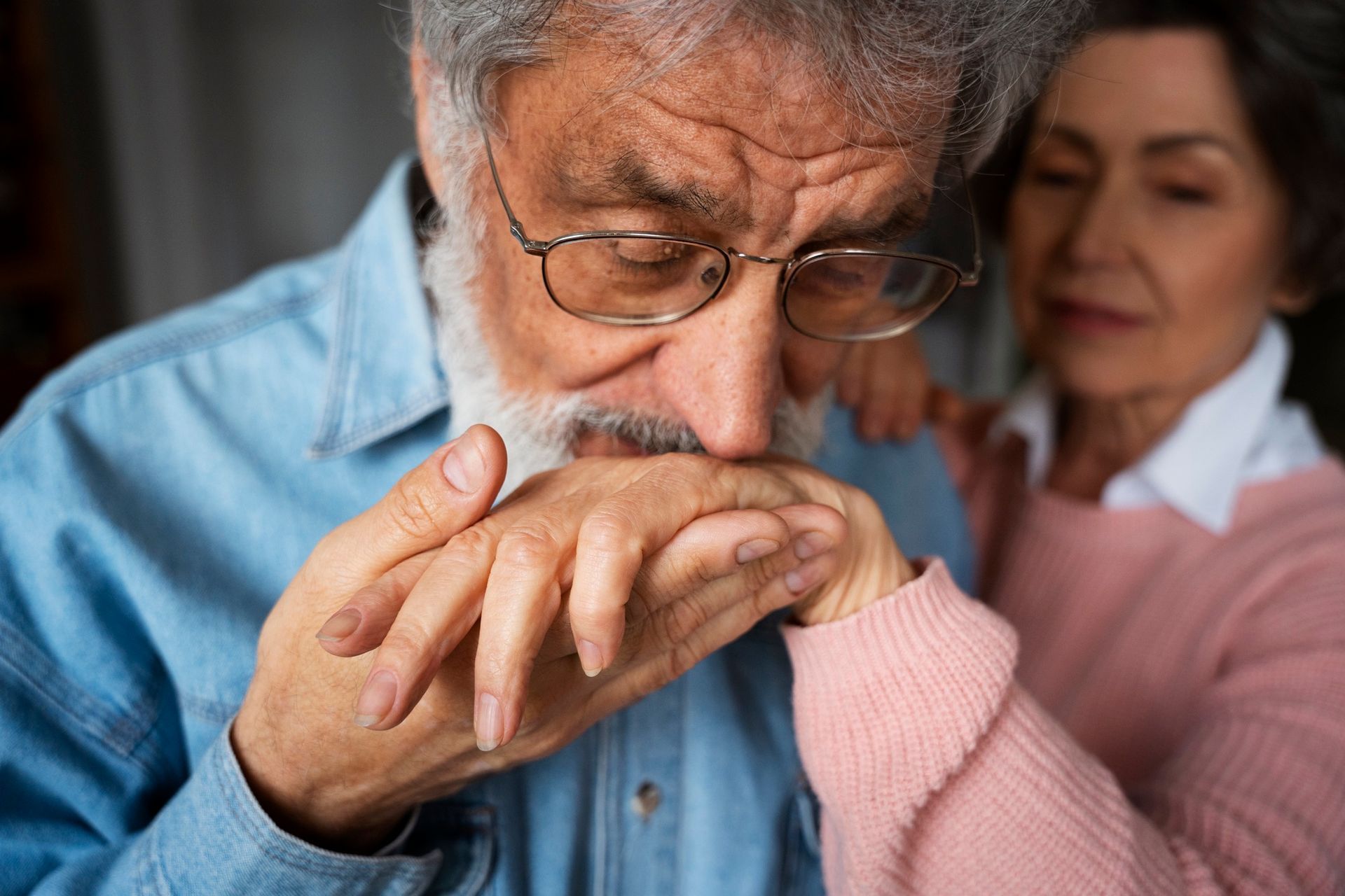 Older man with glasses kissing another person's hand, with a woman in pink smiling in the background
