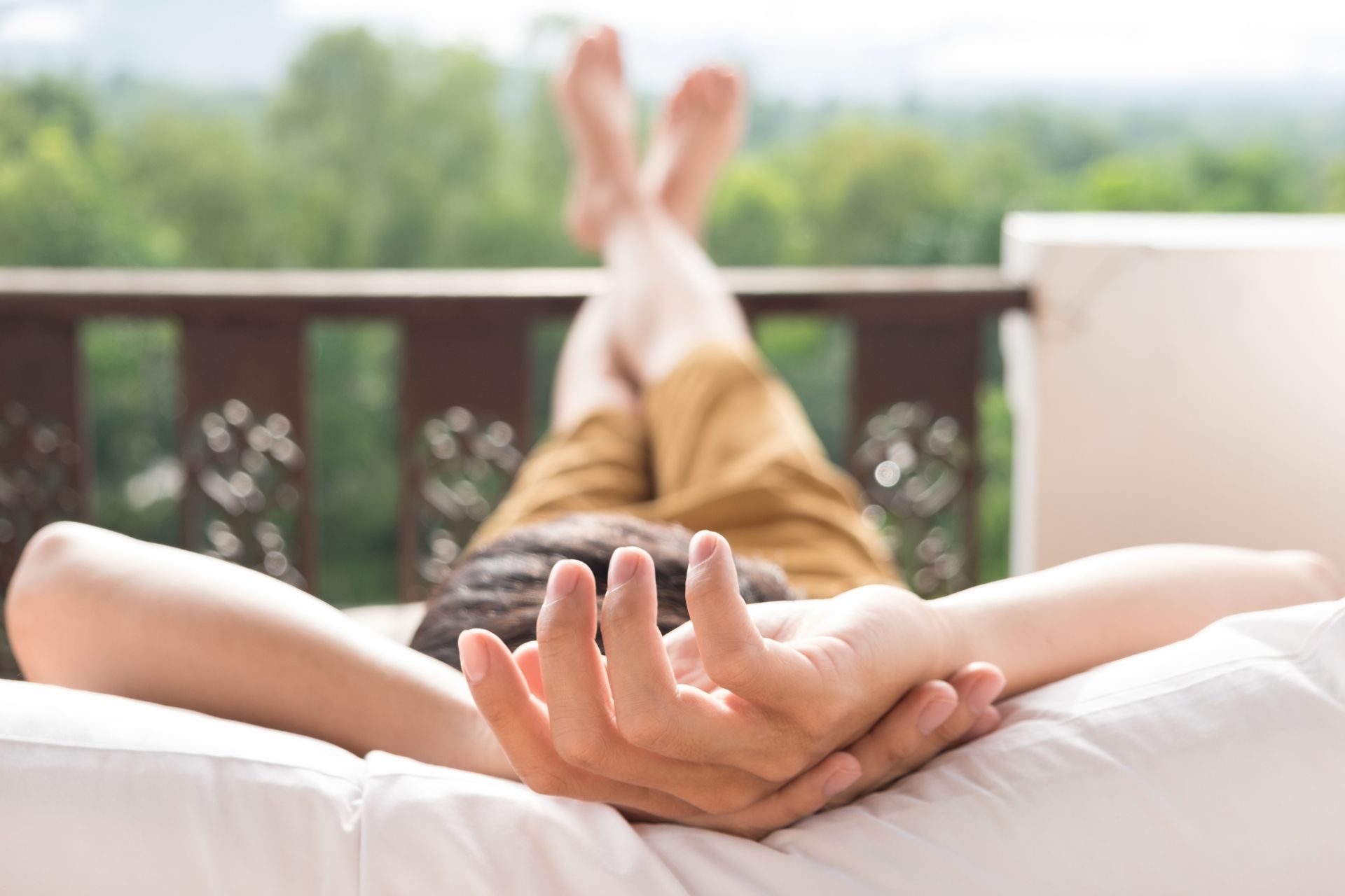 Person lying on a bed with hands clasped in the foreground, outdoors on a balcony overlooking greenery.