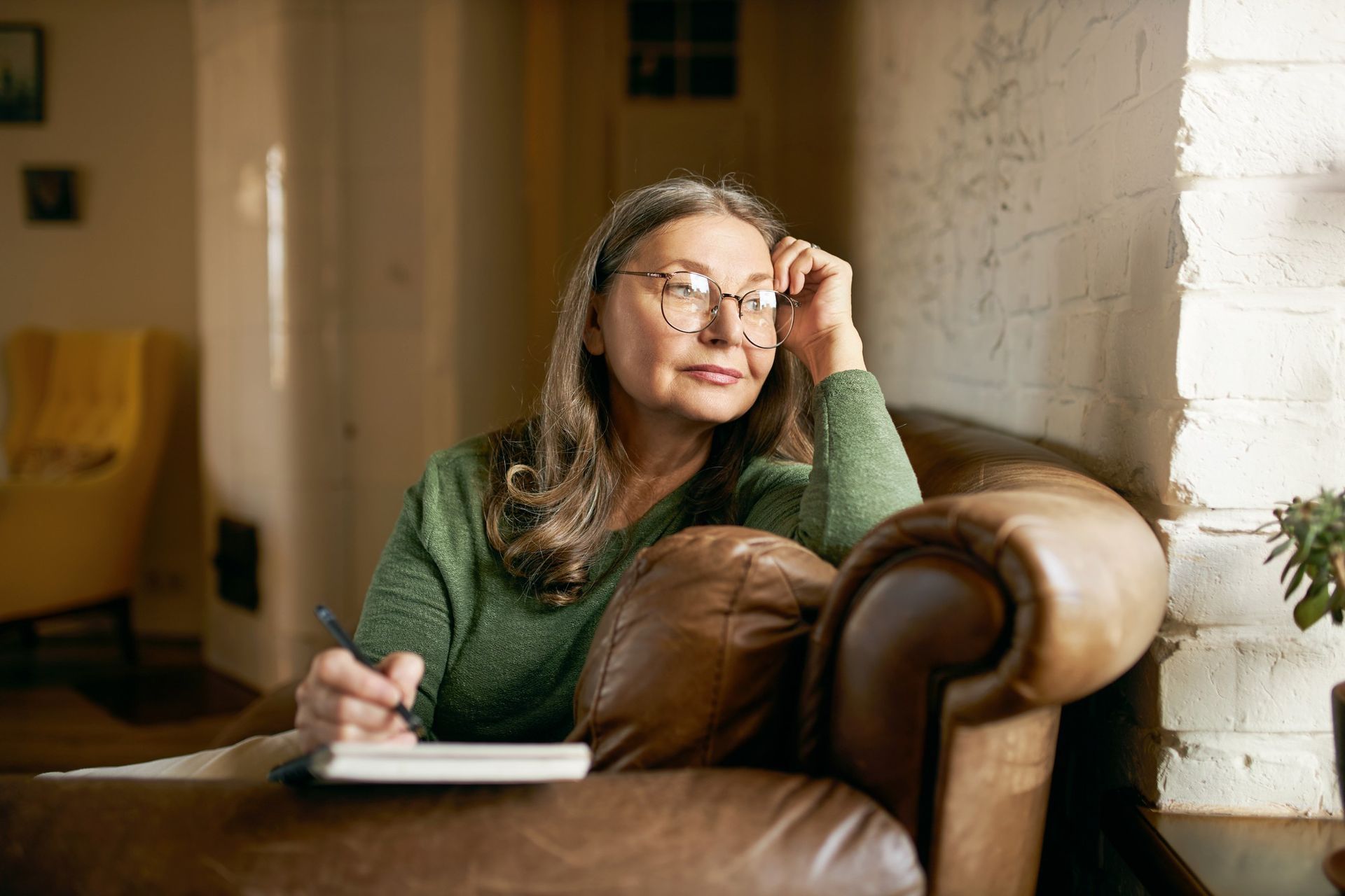 Woman in a green sweater sits on a leather sofa, writing in a notebook and gazing out a window.