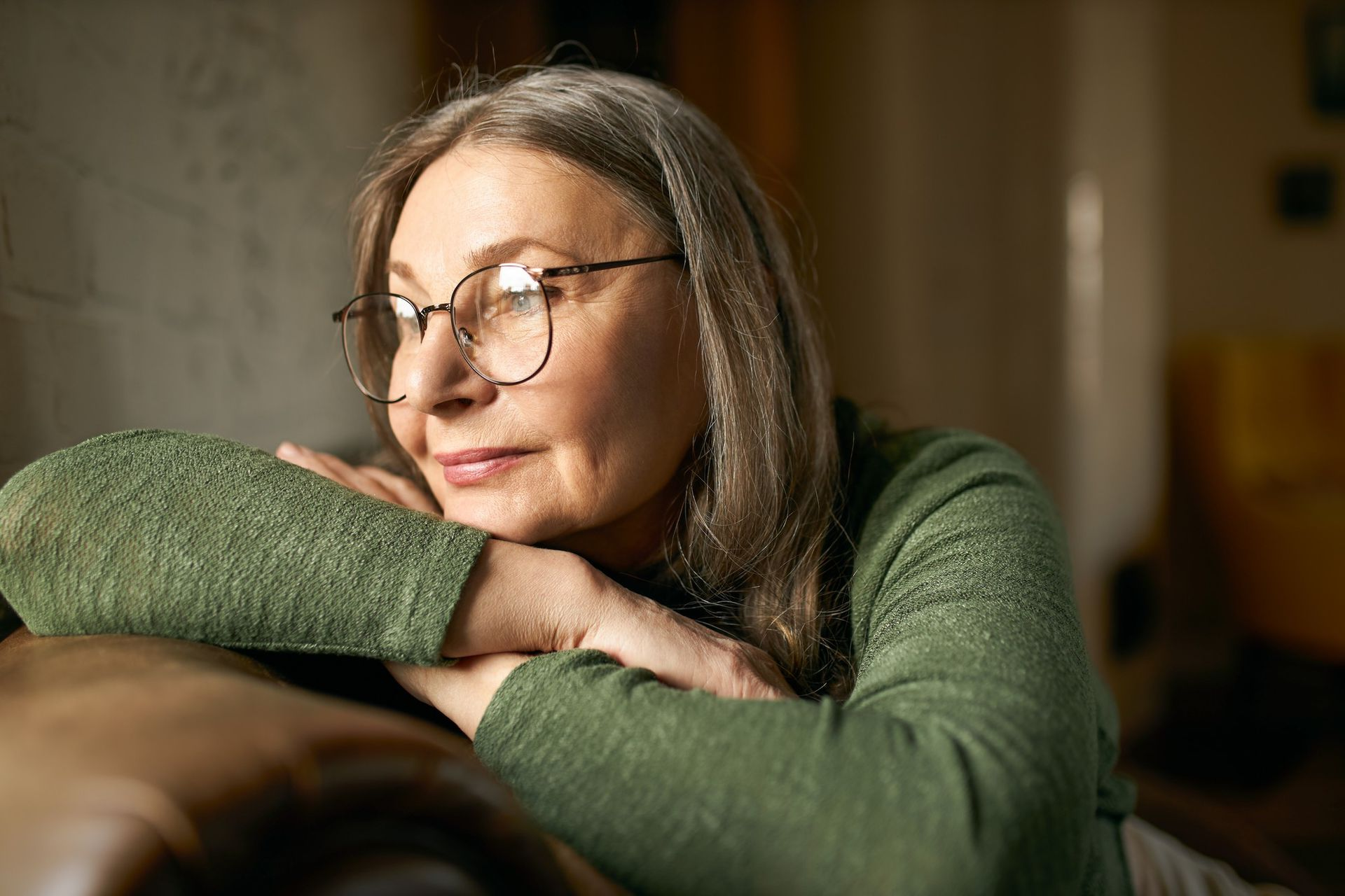 Person in glasses resting their chin on folded arms, wearing a green sweater, looking out a window.