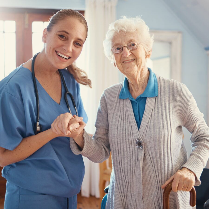 Caregiver in blue scrubs shaking hands with smiling elderly woman holding a cane in a bright home interior