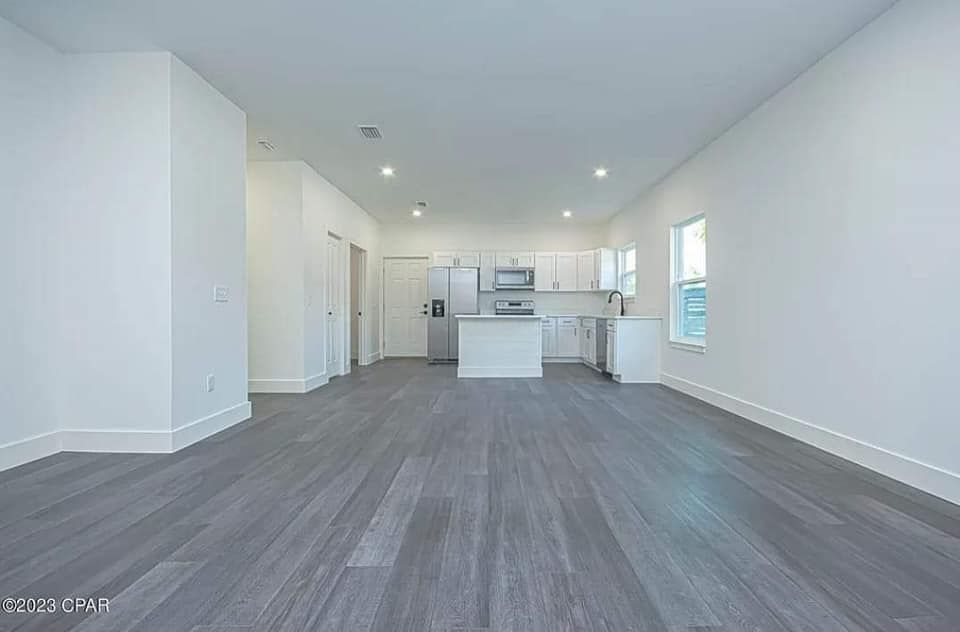 A large empty room with hardwood floors and a kitchen in the background.