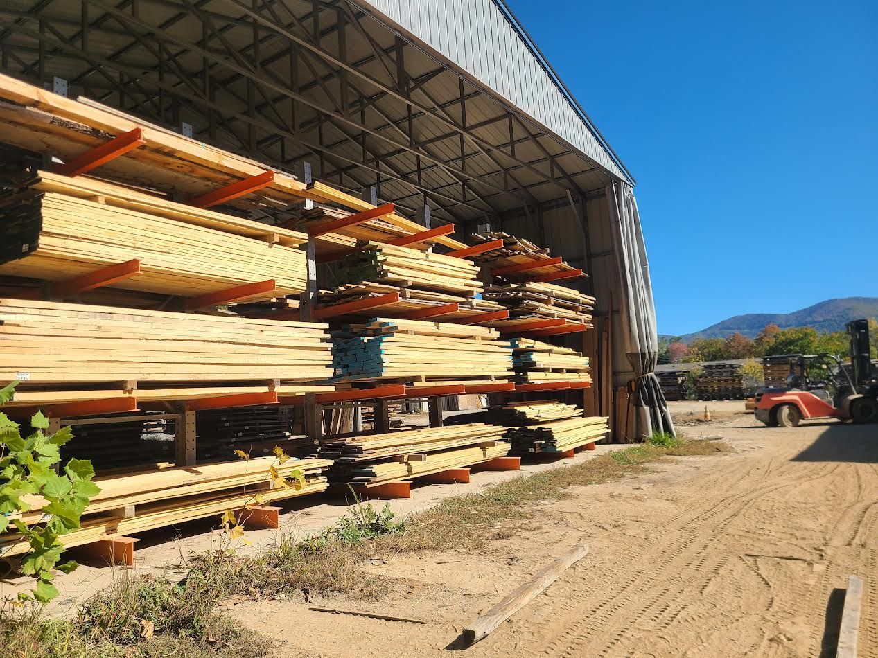 Lumber stacked on racks in a large open-sided shed, under a bright blue sky. A small orange forklift is visible.