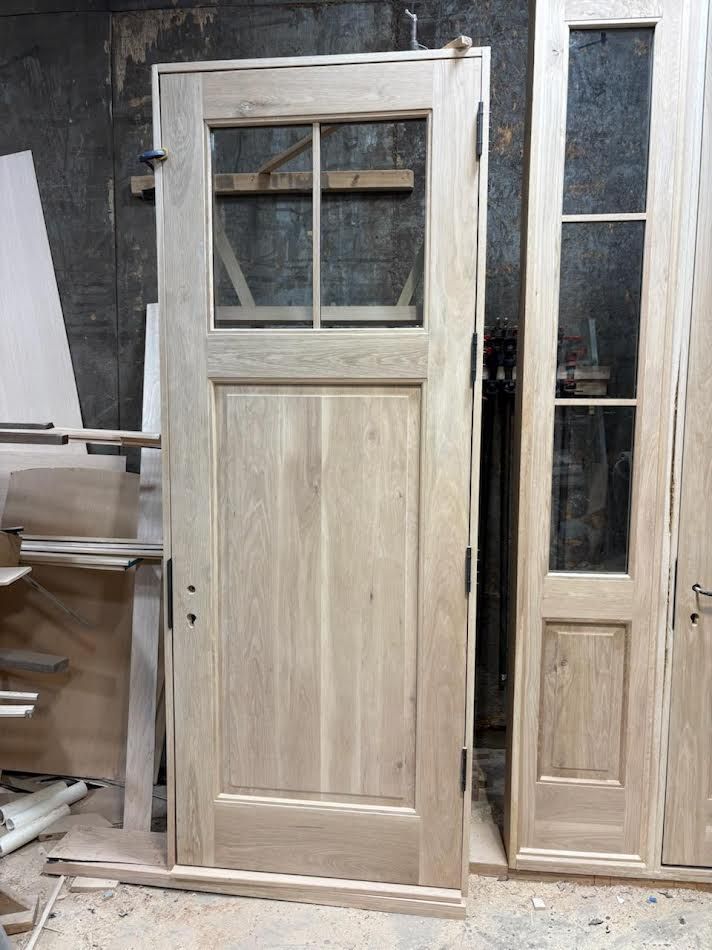 Wooden door with glass panel, unfinished, leaning against a wall in a workshop.