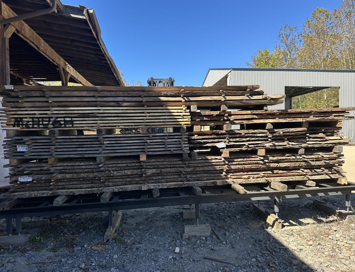 Stacks of weathered wooden planks on pallets outside a barn under a blue sky.