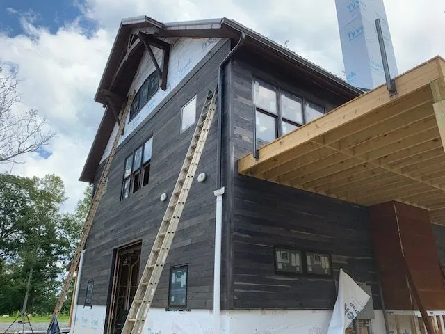 Barn-style house under construction with dark wood siding, windows, and a ladder against the side.